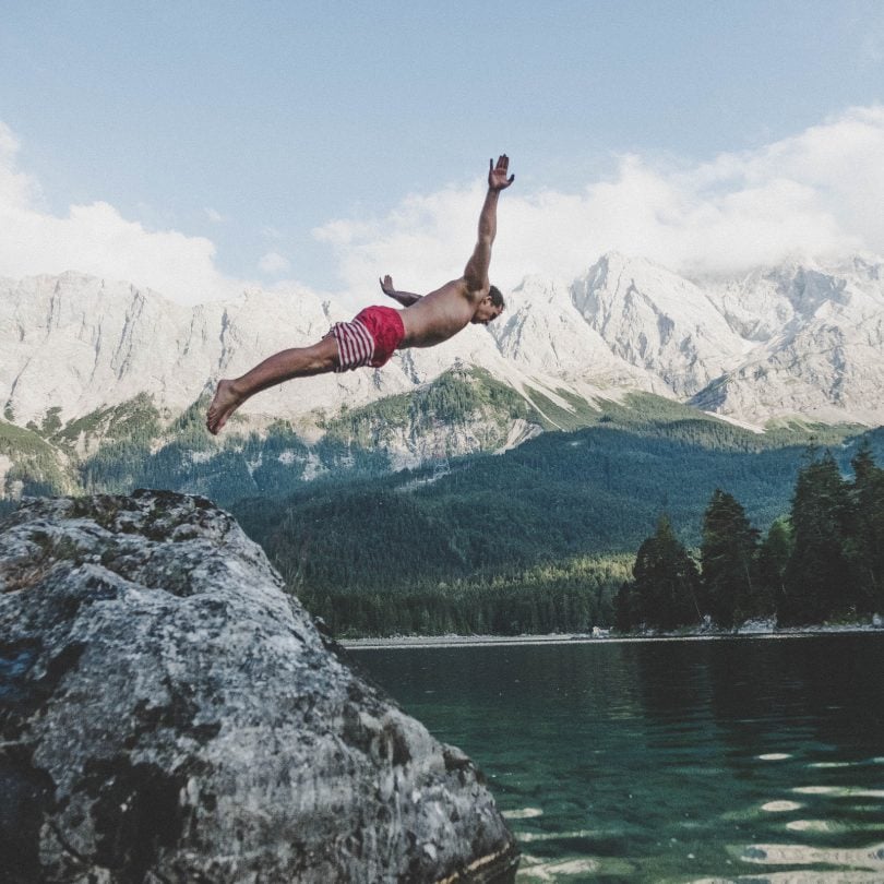 photo of a man jumping into some water near some snow capped mountains
