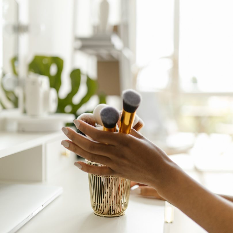 photo of a hand reaching for a make up brush on a shelf, in a cup.